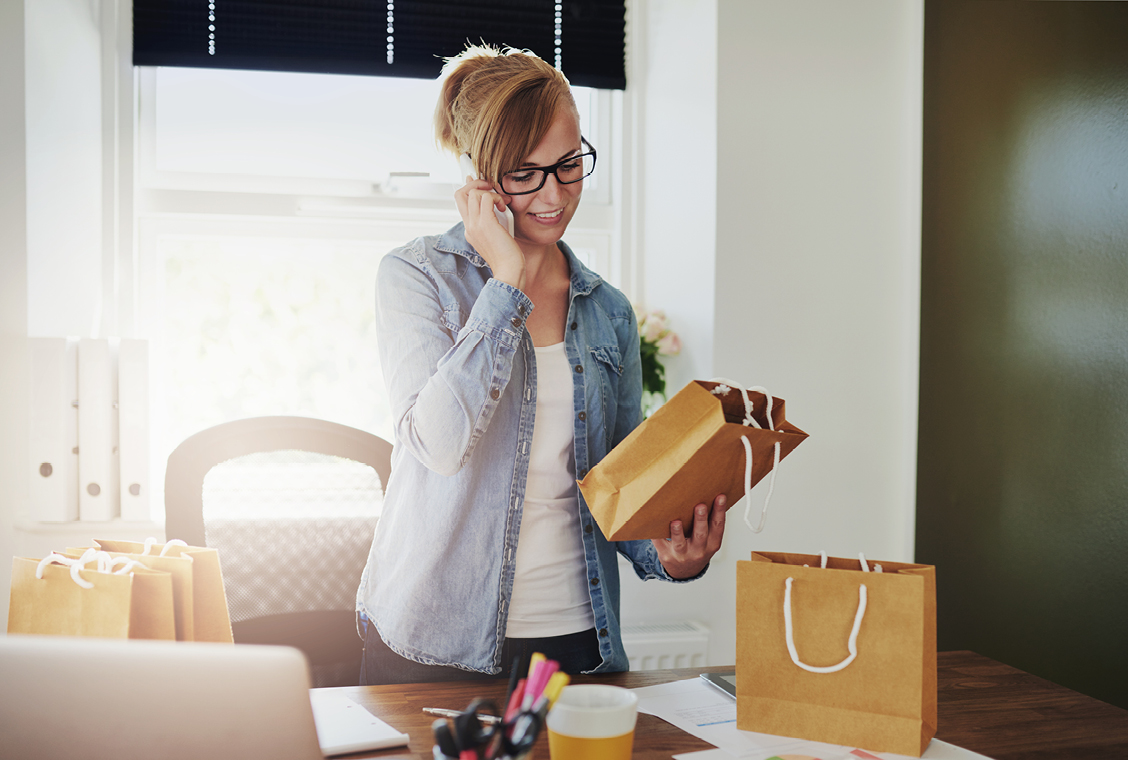 blonde woman in a ponytail, black glasses and a denim shirt on the phone while looking at a brown bag in her hand in her home office.