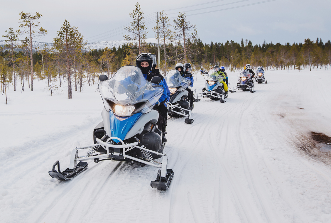 Five snowmobiles with family members riding them drive down a snowy prairie trail in a row.