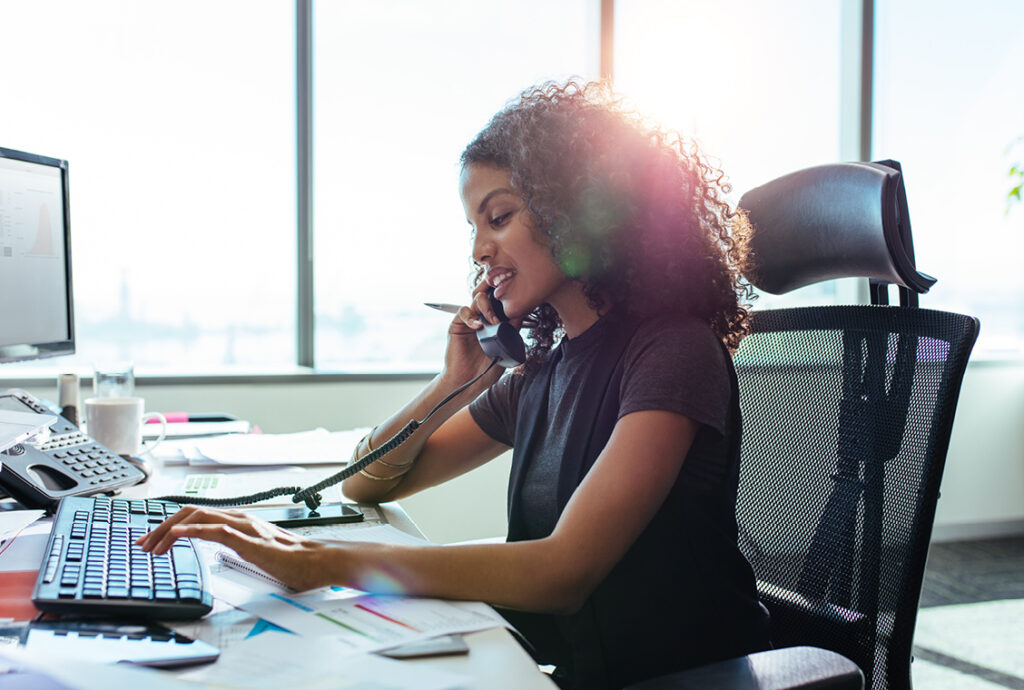 Casually dressed businesswoman with dark brown tightly curled haired talking on the phone at her desk in office.